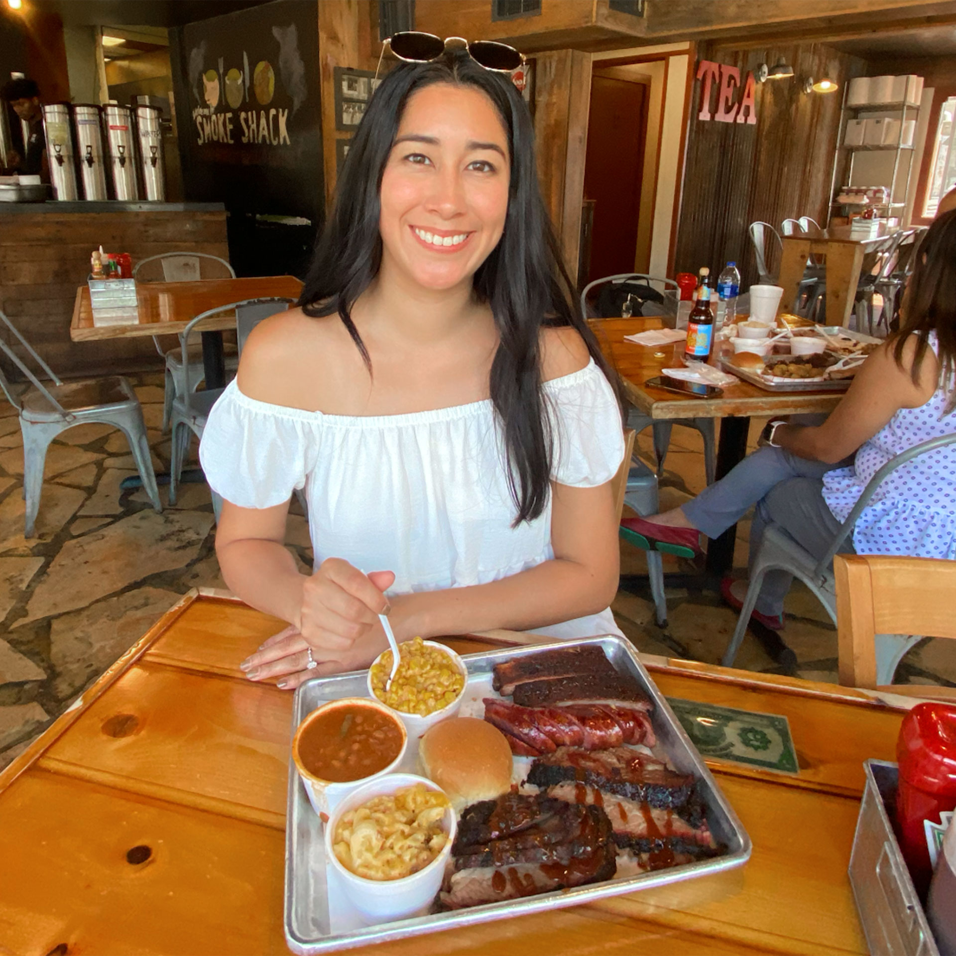 A woman in a white sundress and sunglasses sits at a wooden table with a serving tray containing barbecue dishes and sides.