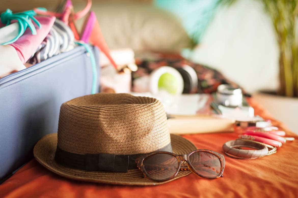 Various items, including a straw fedora, sunglasses, and wooden bangle bracelets, are placed outside of a navy suitcase on an orange bedspread.