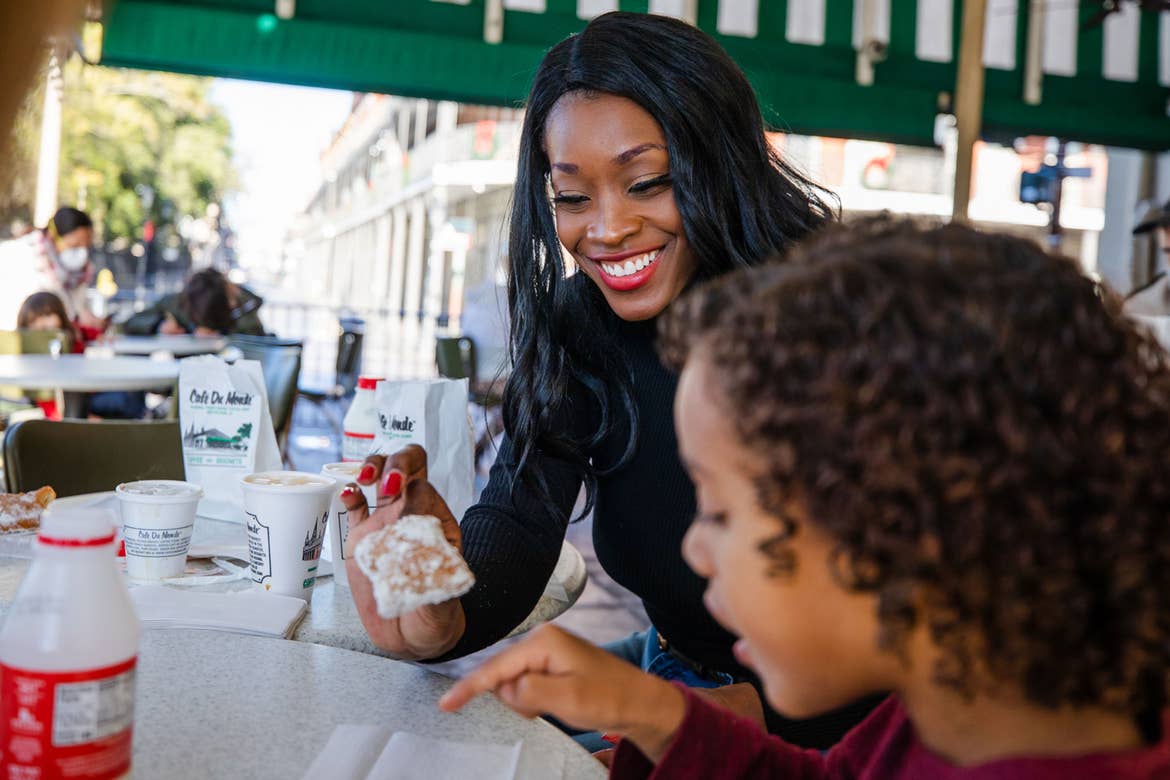 Featured Contributor, Sally Butan of @butanclan (back) and her son, Alex (front), sit under the green canopy outside Cafe Du Monde eating powdery beignets at a white table in New Orleans, Louisiana.
