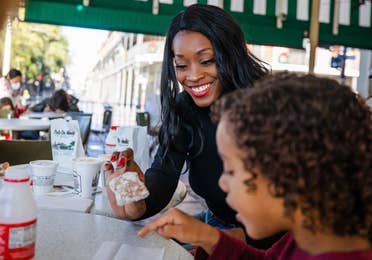 Featured Contributor, Sally Butan of @butanclan (back) and her son, Alex (front), sit under the green canopy outside Cafe Du Monde eating powdery beignets at a white table in New Orleans, Louisiana.