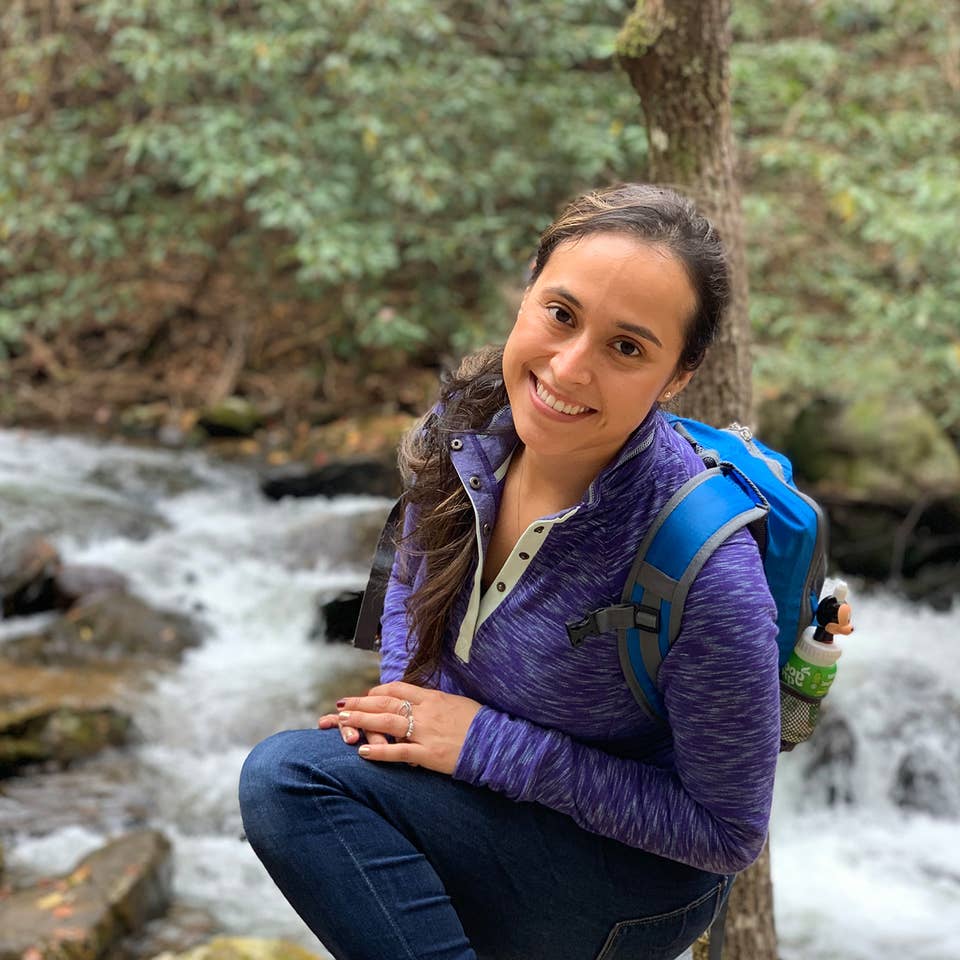 Featured author, Andrea Beltran, poses near a creek wearing a blue hiking backpack and purple pull-up sweater.