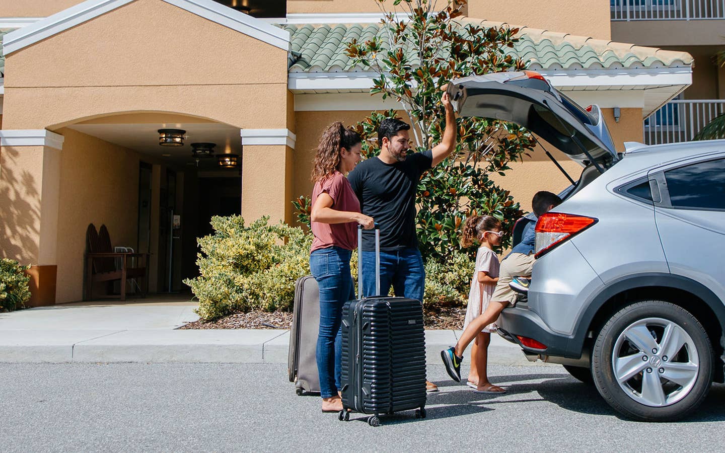 A woman, man and young boy and girl unpack a silver vehicle outside of a resort.