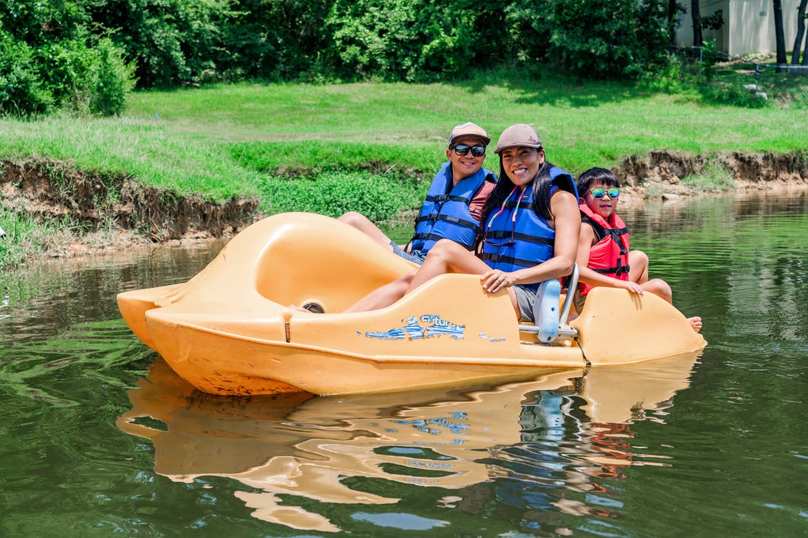 An Asian Male (left), female (middle) and young child (right) wear blue life vests, sunglasses and various hats while riding on a yellow paddleboat in a lake.