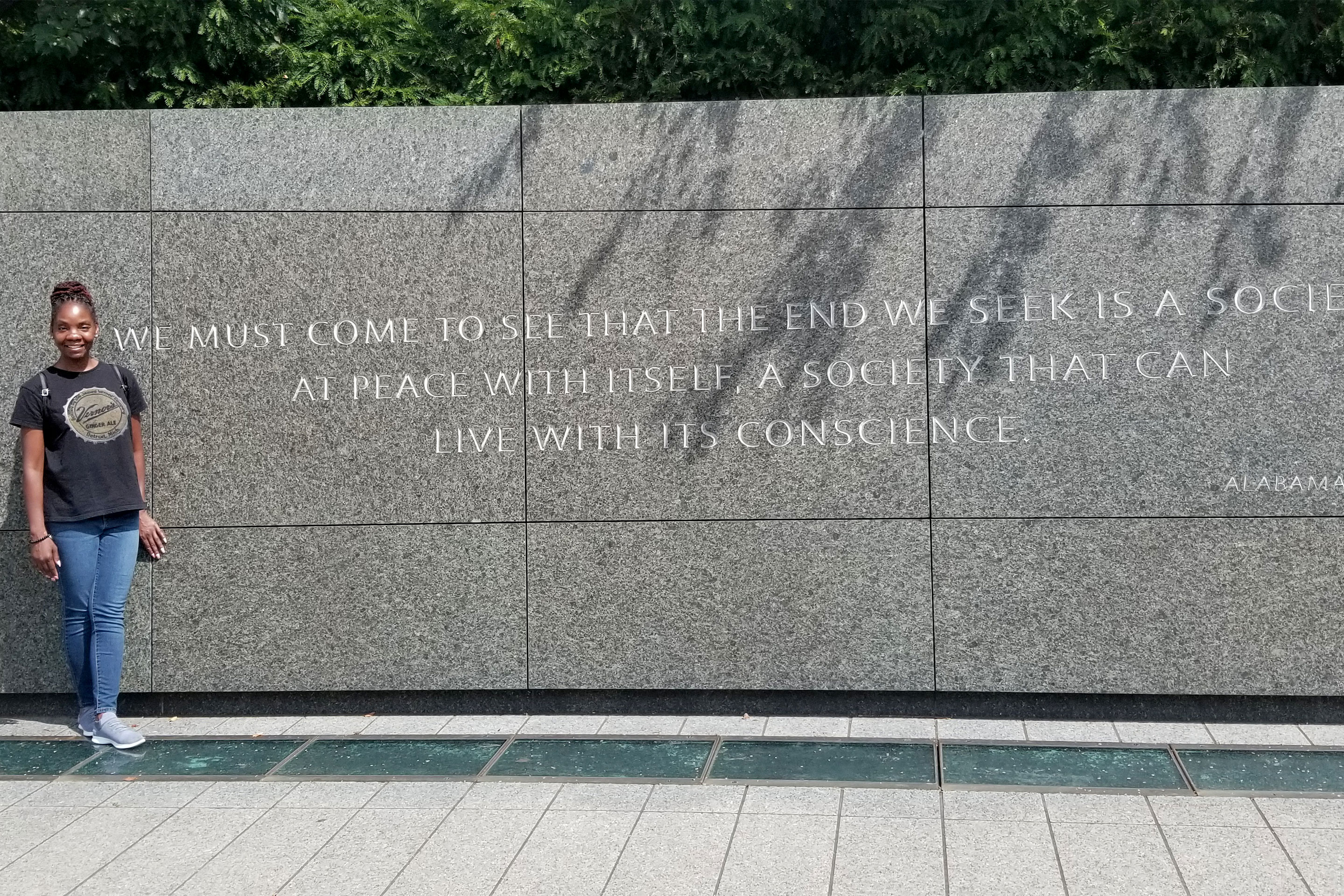 A woman in a black t-shirt, and jeans stands next to a memorial wall with a Martin Luther King Quote.