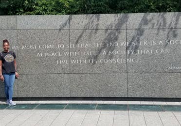 A woman in a black t-shirt, and jeans stands next to a memorial wall with a Martin Luther King Quote