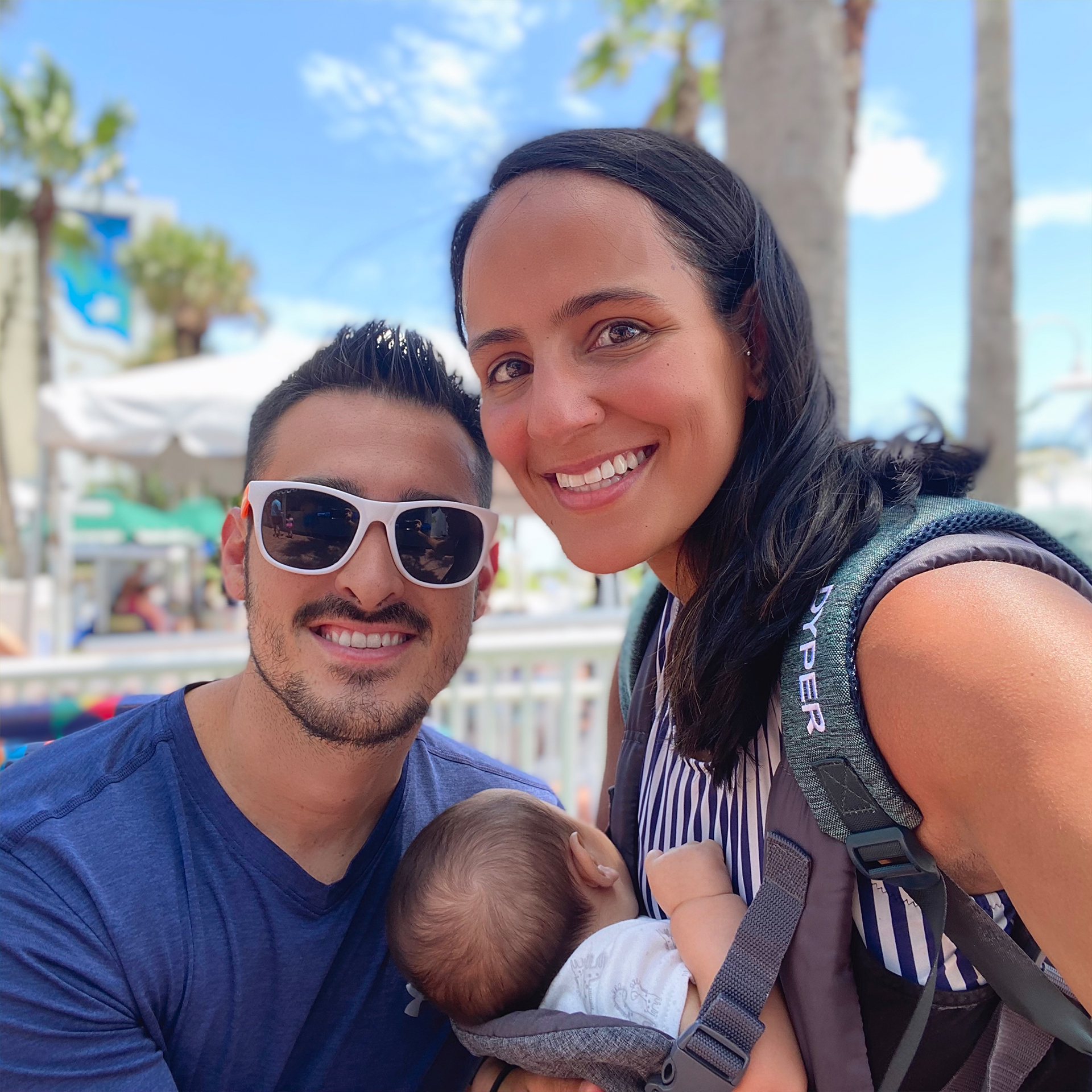 A man in a wheelchair and a woman standing while carrying a baby outdoors.