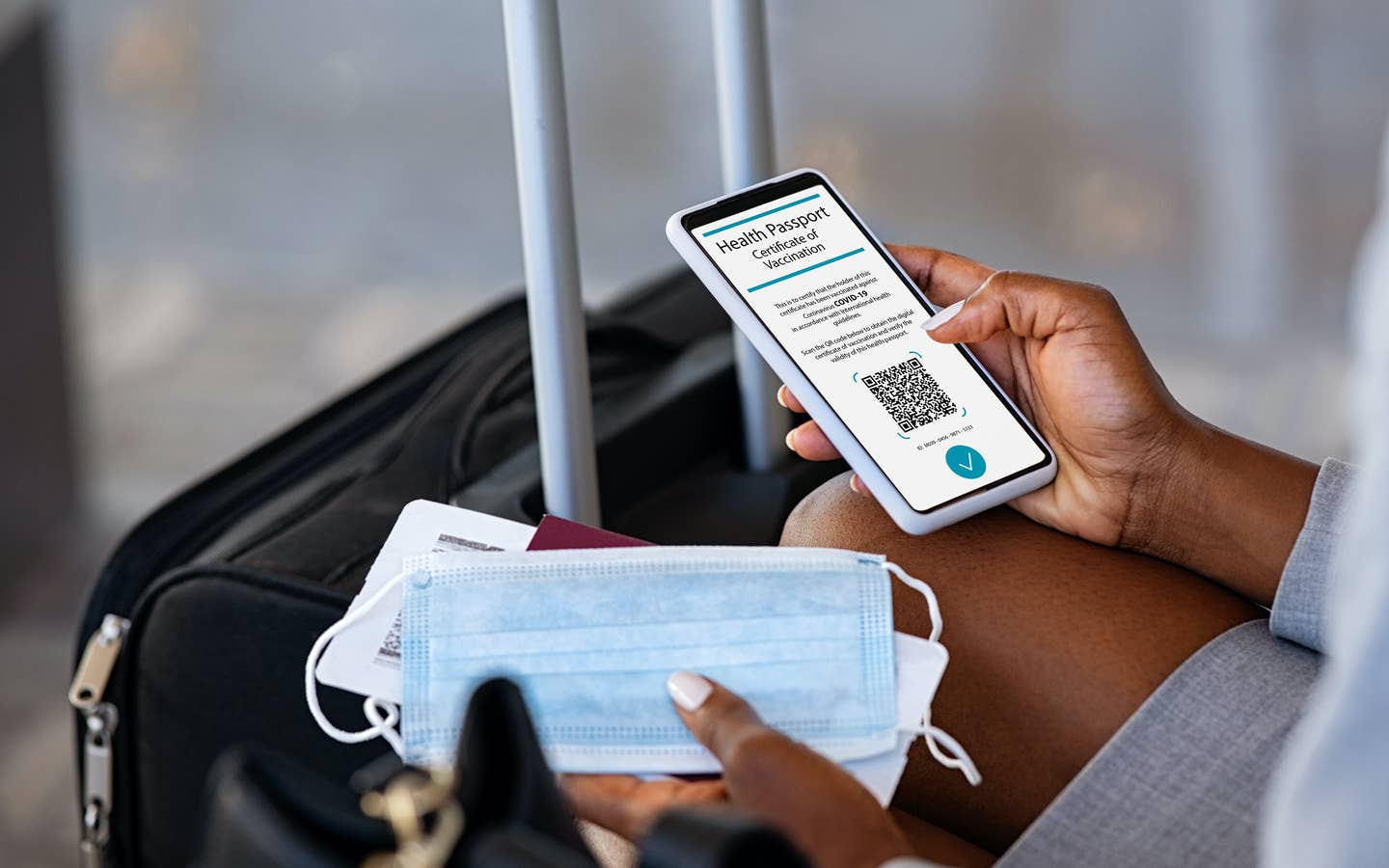 Traveler pulling out their cell phone to look at their digital health pass/vaccine passport, with a mask in their hand and luggage in front of them.