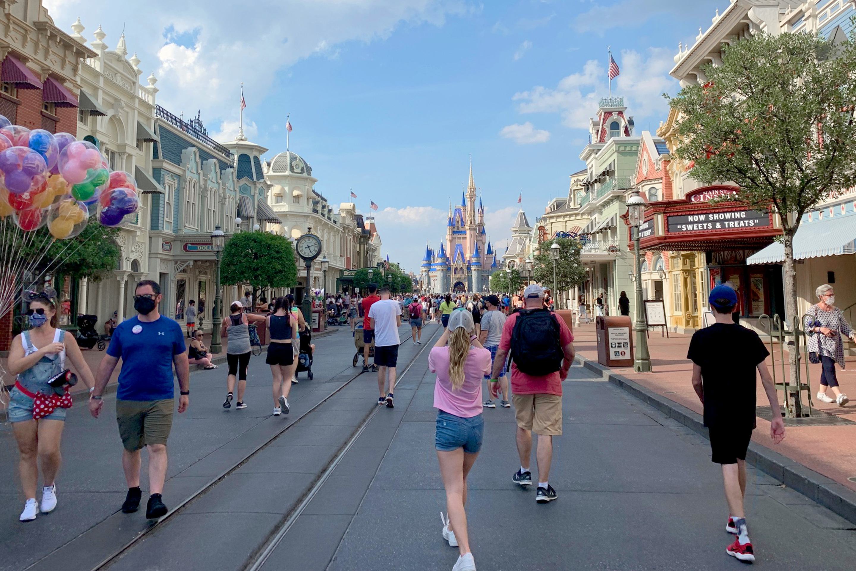 Several guests wearing safety masks walk up and down Main Street USA at the Magic Kingdom at Walt Disney World Resort as Cinderella's Castle stands in the distance under a cloudy blue sky.