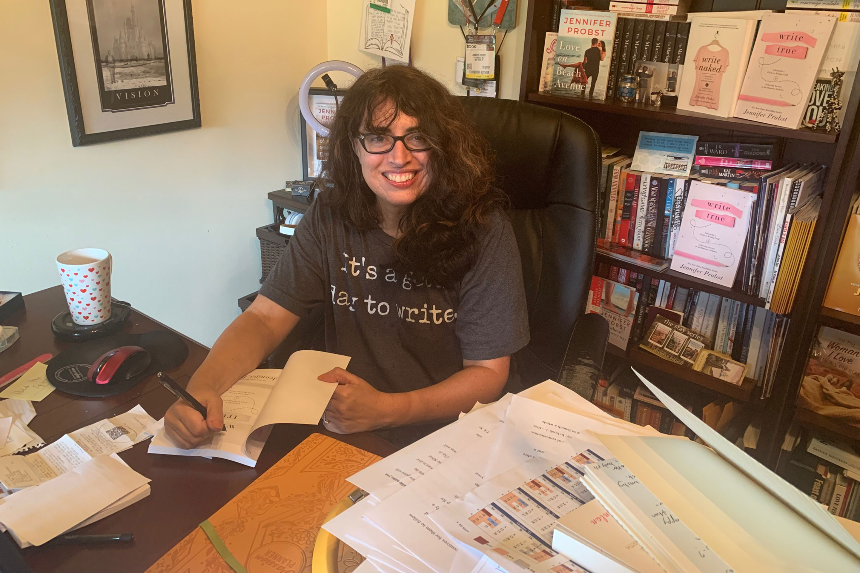 Author, Jennifer Probst, wears her glasses and a grey shirt while writing a book on her desktop.