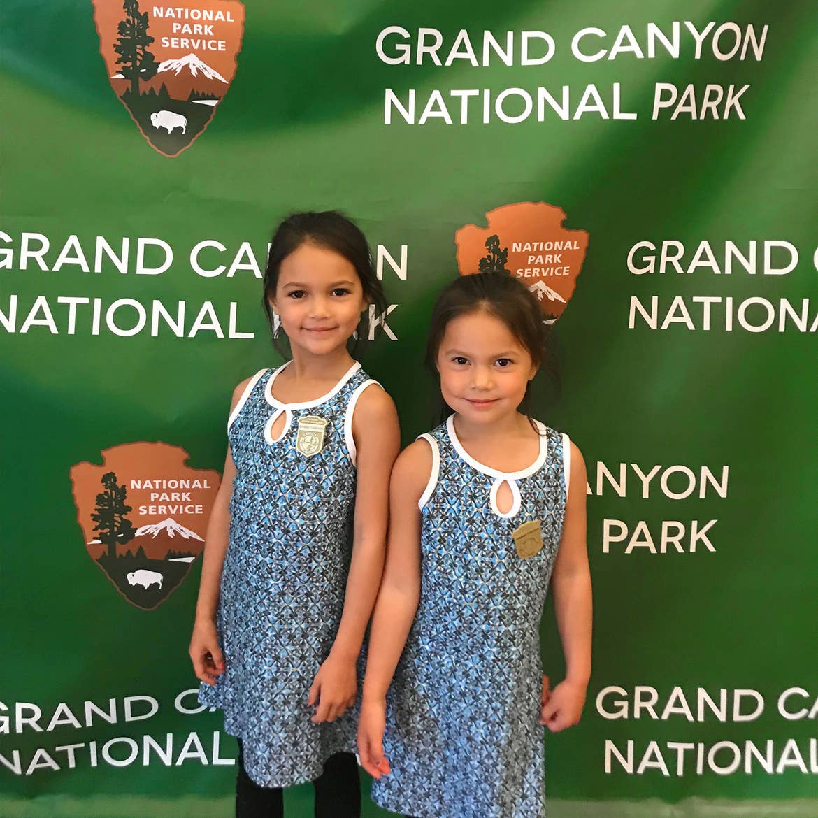 Two young girls in matching blue and white dresses wear two Junior Park Ranger badges in front of a green backdrop reading, 'Grand Canyon National Park'.