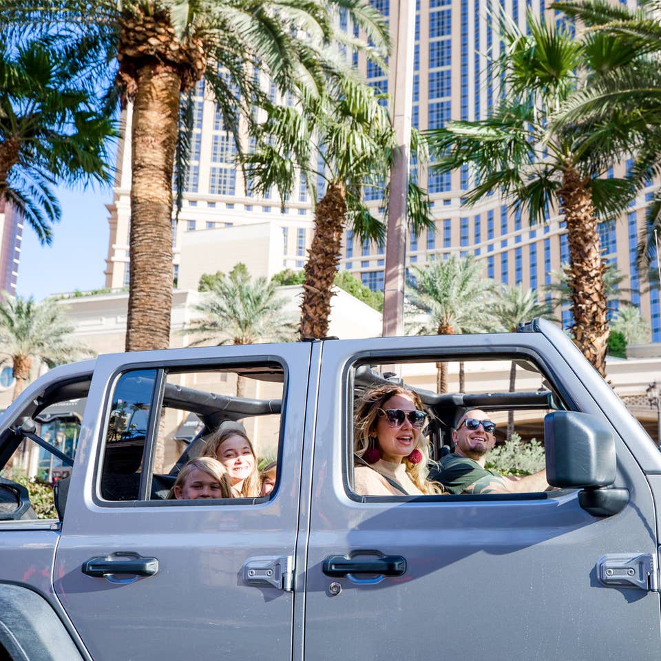 A man drives a silver Jeep Wrangler with one female passenger and three children in the back though the Las Vegas strip with the roof shell off.
