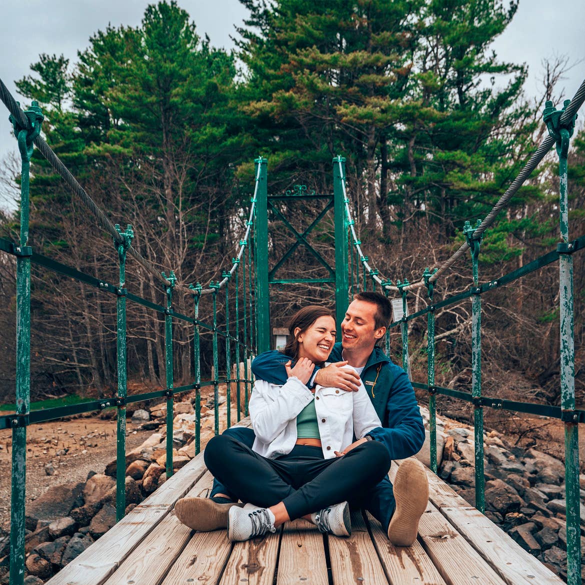 A man wearing a dark hoodie embraces a woman in a white jacket and black leggings as they sit on a suspension bridge surrounded by green trees.