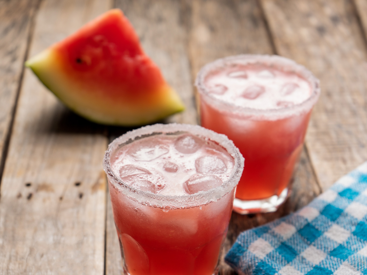 Two glasses of Melon Margaritas sit on a wooden plank table in front of a watermelon wedge.
