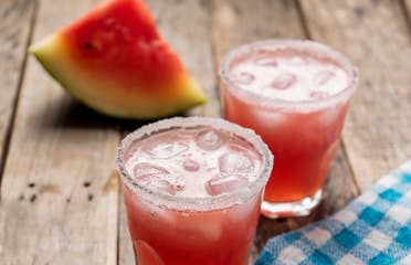 Two glasses of Melon Margaritas sit on a wooden plank table in front of a watermelon wedge.