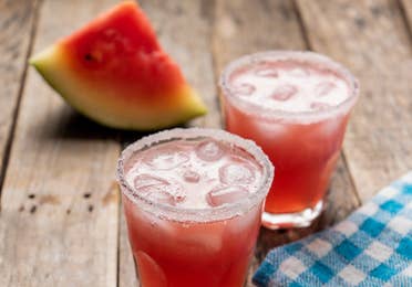 Two glasses of Melon Margaritas sit on a wooden plank table in front of a watermelon wedge.