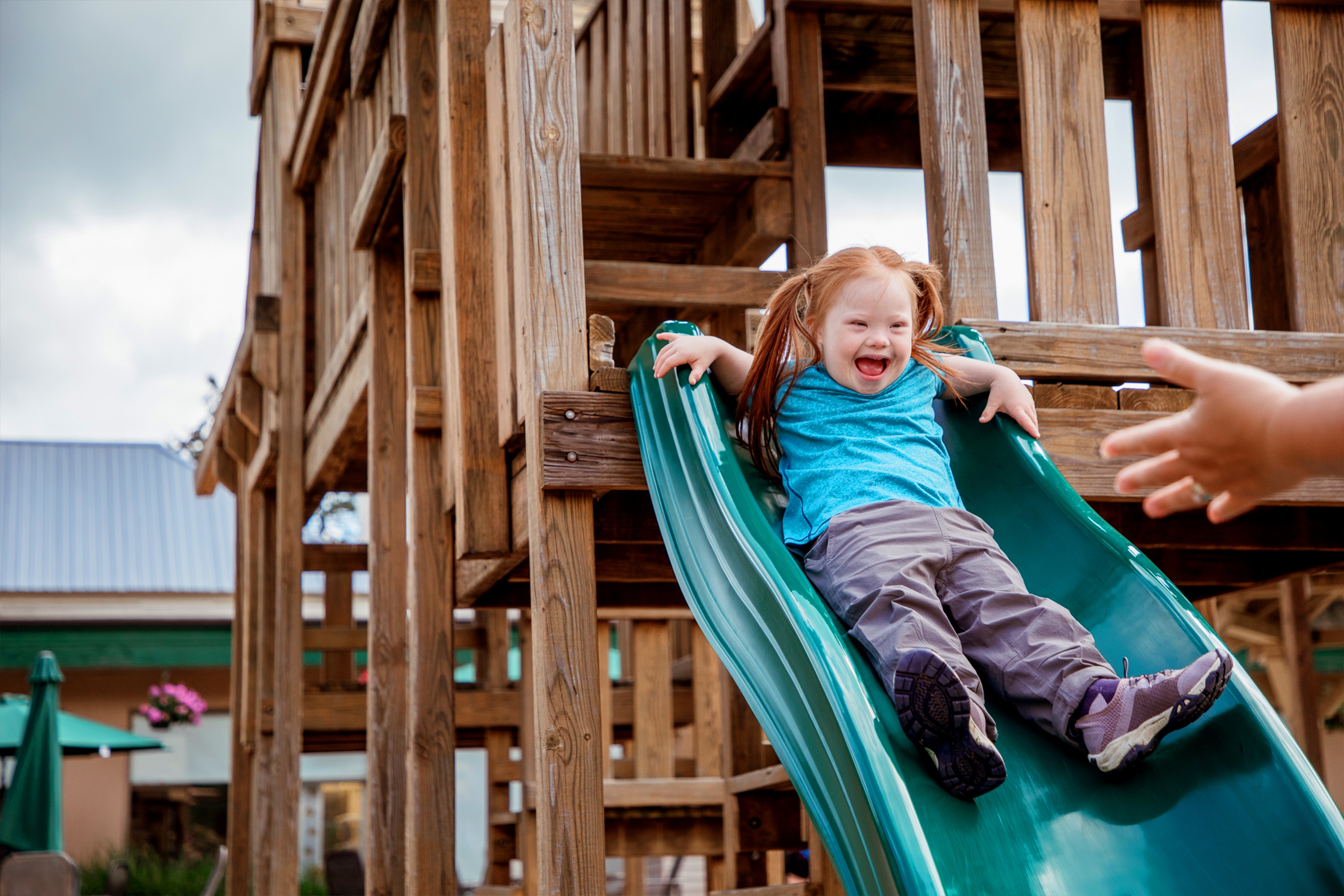 A young, caucasian girl with red hair, an aqua shirt and khaki-colored pants slides down a wooden playground slide outdoors.