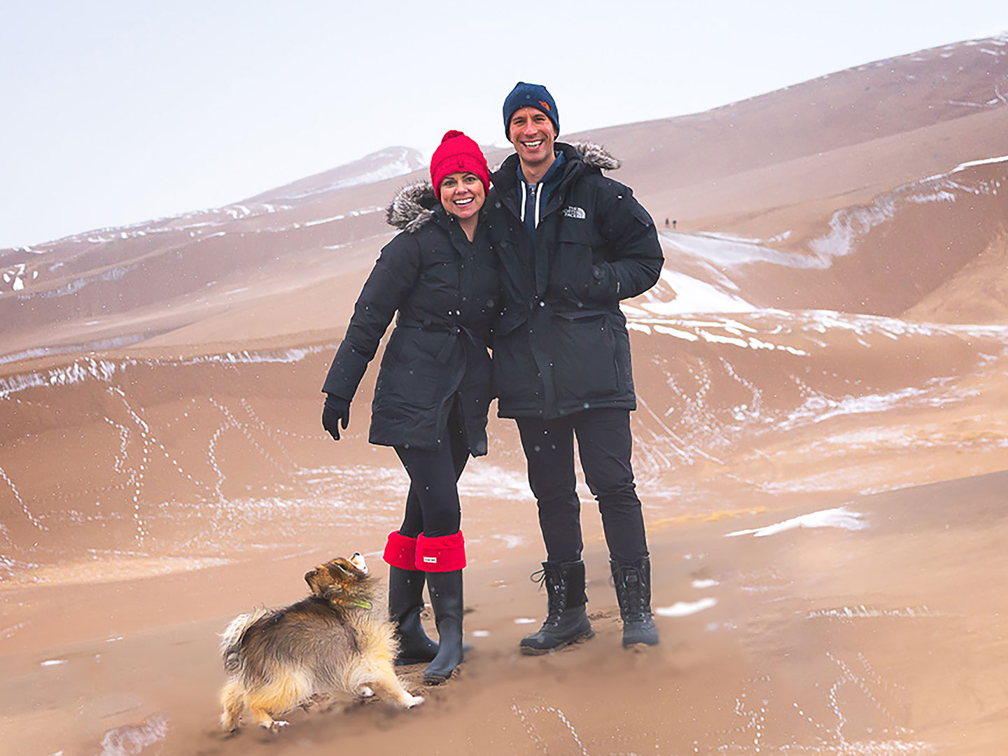 Authors, Lauren Layne and Anthony LeDonne, stand in winter apparel with Bailey the Pomeranian at the Great Sand Dunes National Park and Preserve.
