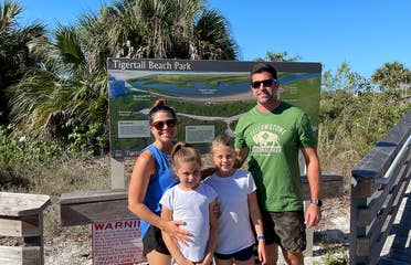 A caucasian family of four stand in front of a trail map with Palm Trees in the back.