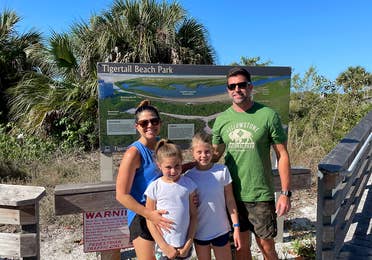 A caucasian family of four stand in front of a trail map with Palm Trees in the back.