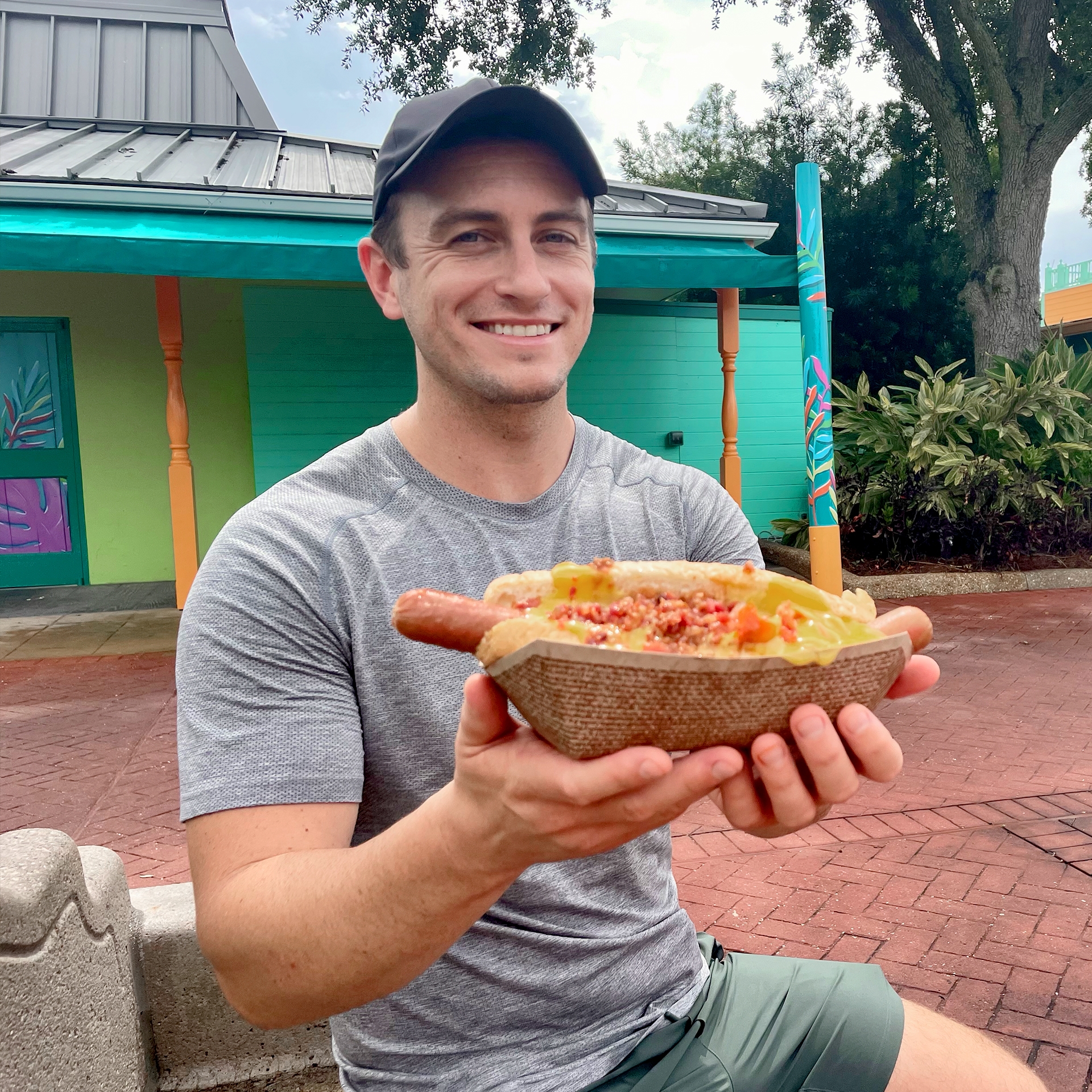 A Caucasian male wearing a black baseball cap, grey shirt and khaki shorts holds a cheese-covered hot dog while seated in SeaWorld Orlando.
