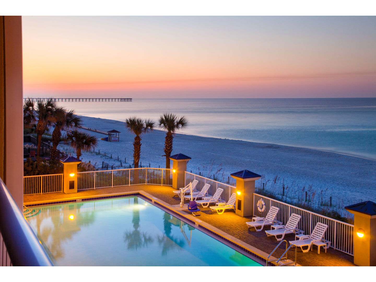 Beachfront resort pool overlooking the ocean at sunset with palm trees.