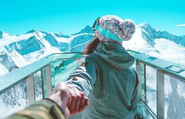 A woman holds a man's hand while wearing winter apparel and ski goggles looking off at snowcapped mountains under a blue sky.