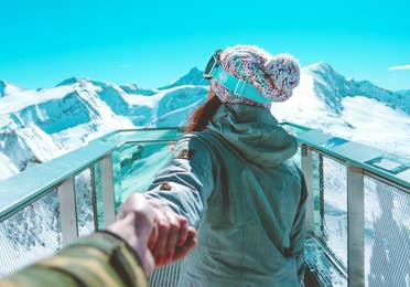 A woman holds a man's hand while wearing winter apparel and ski goggles looking off at snowcapped mountains under a blue sky.