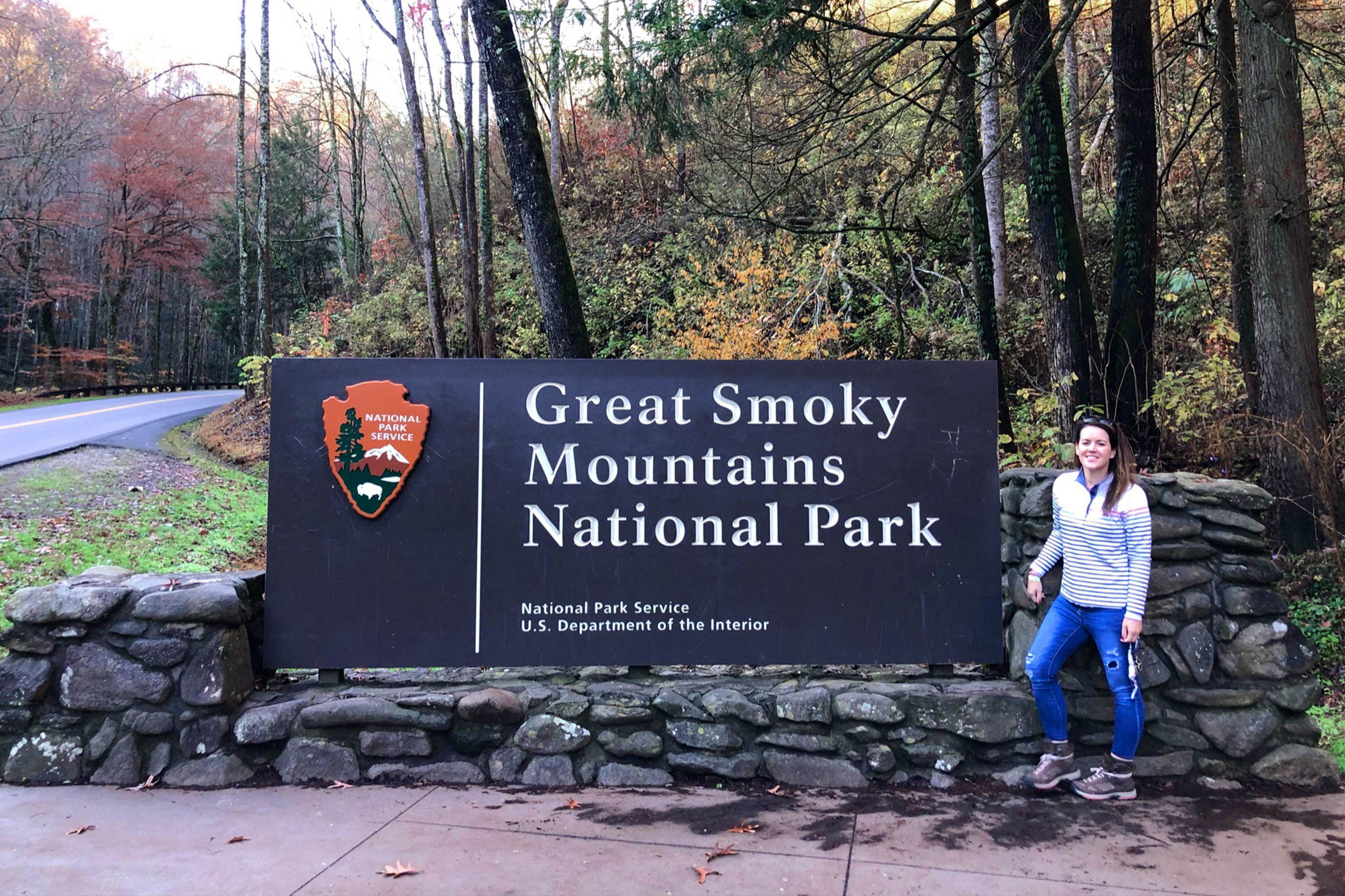 Featured Contributor, Jennifer C. Harmon, poses with a sign that reads, 'Great Smoky Mountains National Park' wearing a white pullover and jeans.