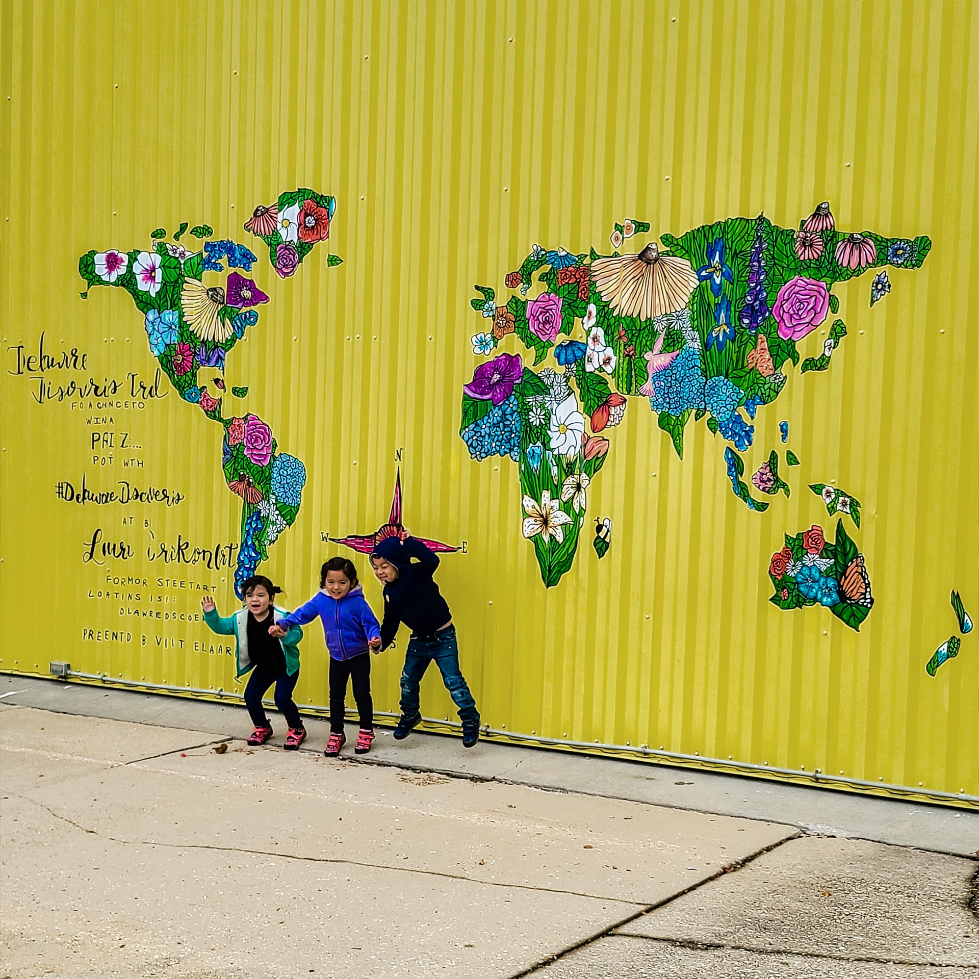 Three Asian Pacific Islander toddlers (left to right: Two girls and a boy) wear long-sleeve shirts and pants in front of a galvanized yellow wall with a mural of the world map with floral patterns.