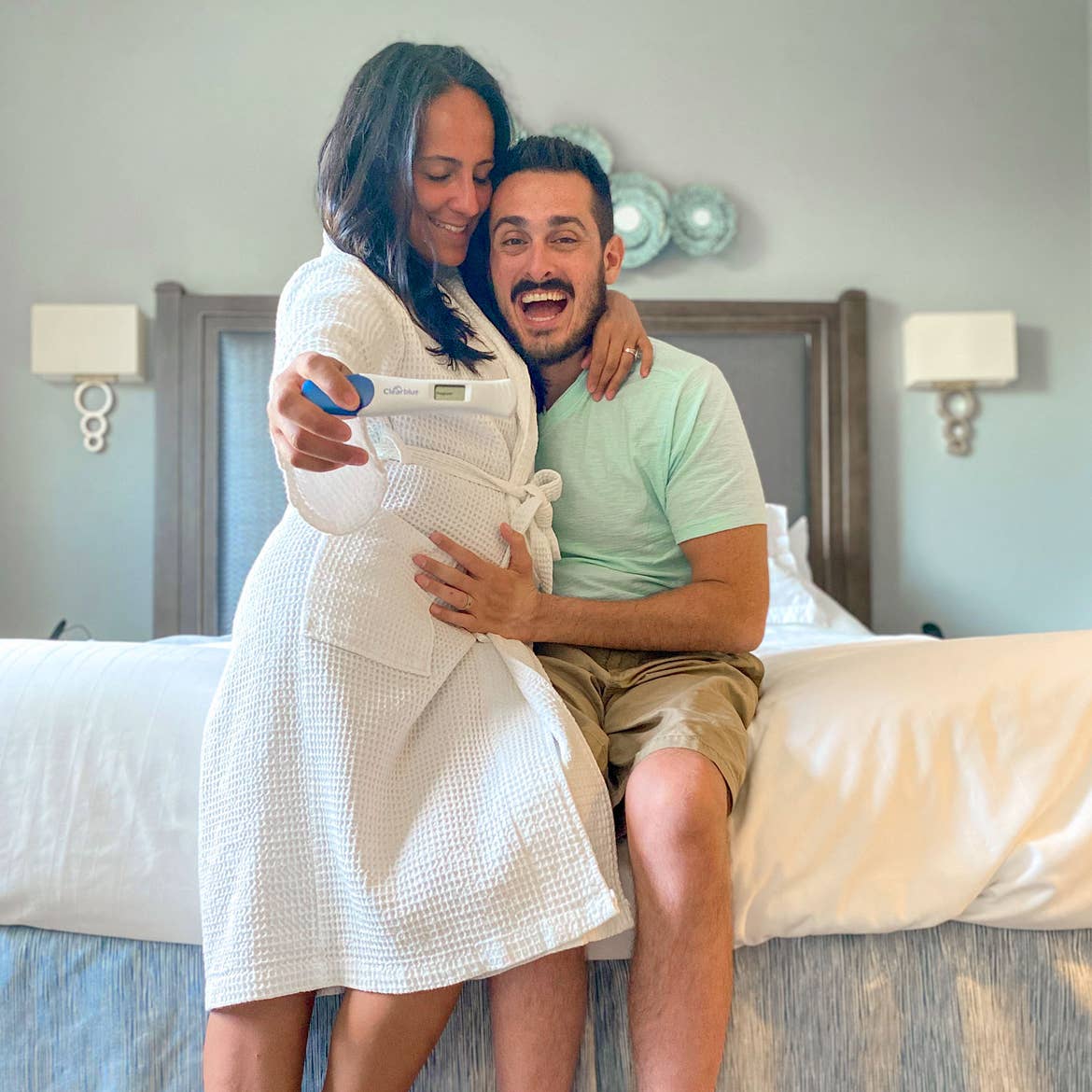 Featured Contributor, Danny Pitaluga (right) smiles as he hugs his wife, Val (left), as she holds a positive pregnancy test in a white robe.