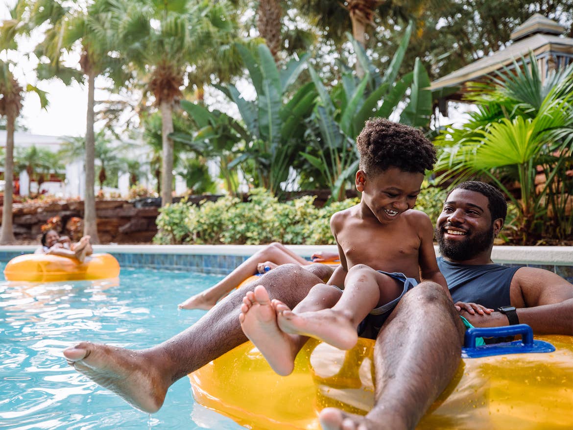 A young boy sits on a man's lap as they float on a yellow inner-tube on an outdoor lazy river.