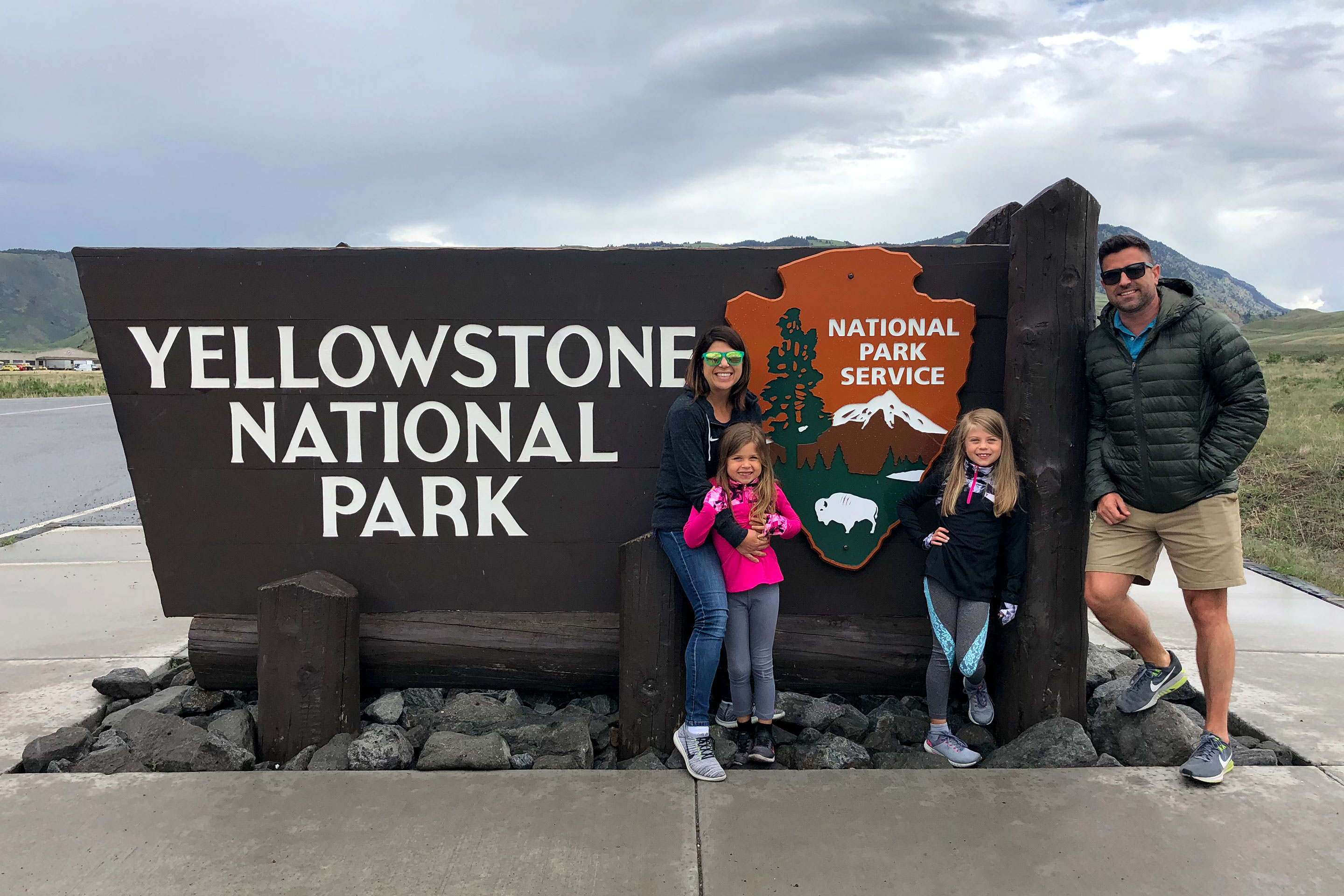 Author, Chris Johnston (far-left), with her husband, Josh (far-right), and daughters, Kyndall (front-right), and Kyler (front-left) pose with a sign that reads, 'Yellowstone National Park, National Park Service.'