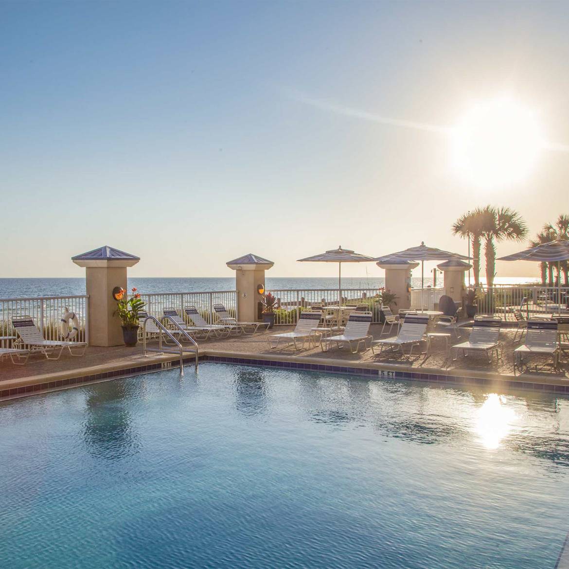 Sun shines over beachfront pool lined with lounge chairs.