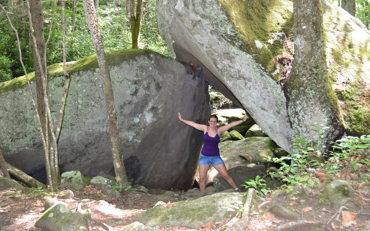 A woman wearing a purple tank top, jean shorts and glasses poses underneath a pair of wedged rocks in Gatlinburg, TN.