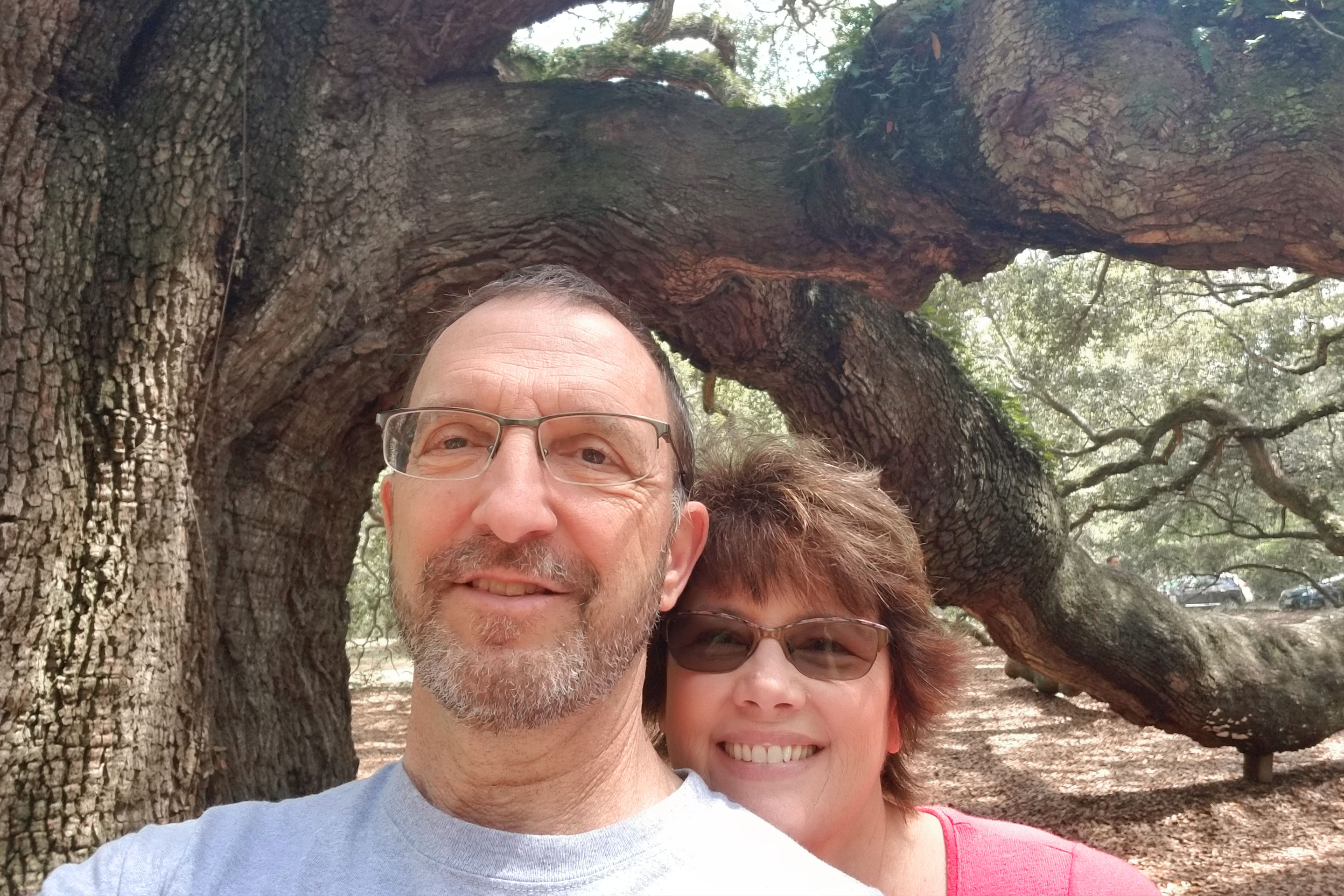 A woman wearing glasses and a pink t-shirt stands behind a man in reading glasses and white shirt next to a moss tree on a walking trail.