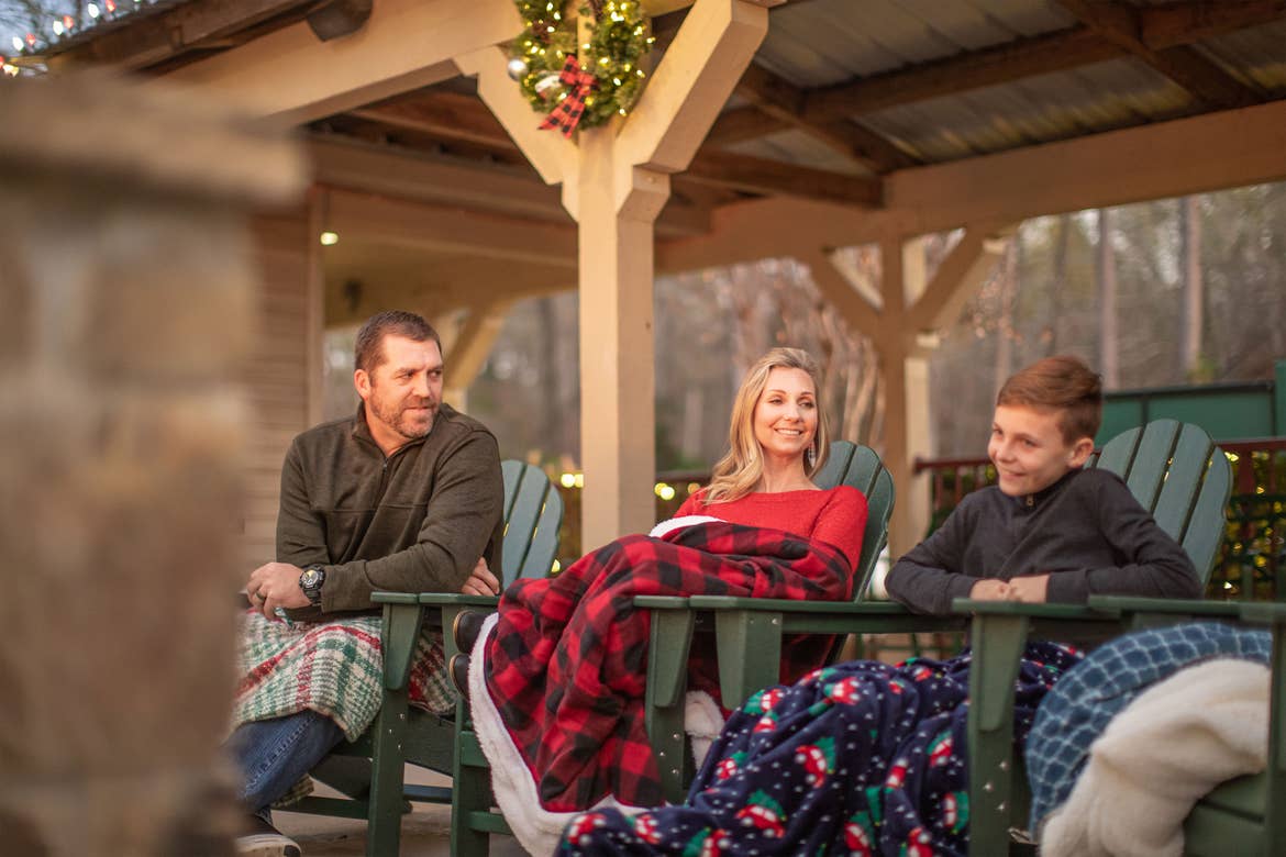 Author, Amanda Nall (middle) sits outdoors with her husband (left) and son (right) wearing cozy throw blankets outdoors.