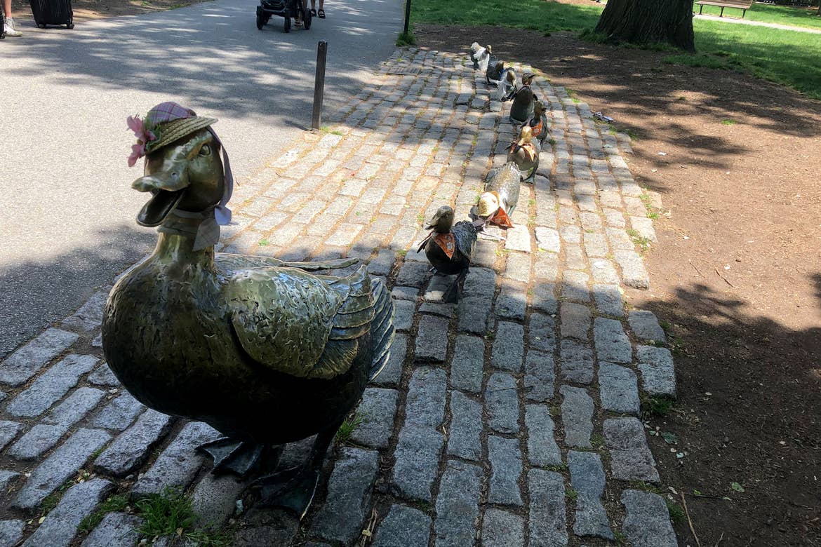 Several bronze statues of a duck and her ducklings wear bonnets on a brick paved walkway.