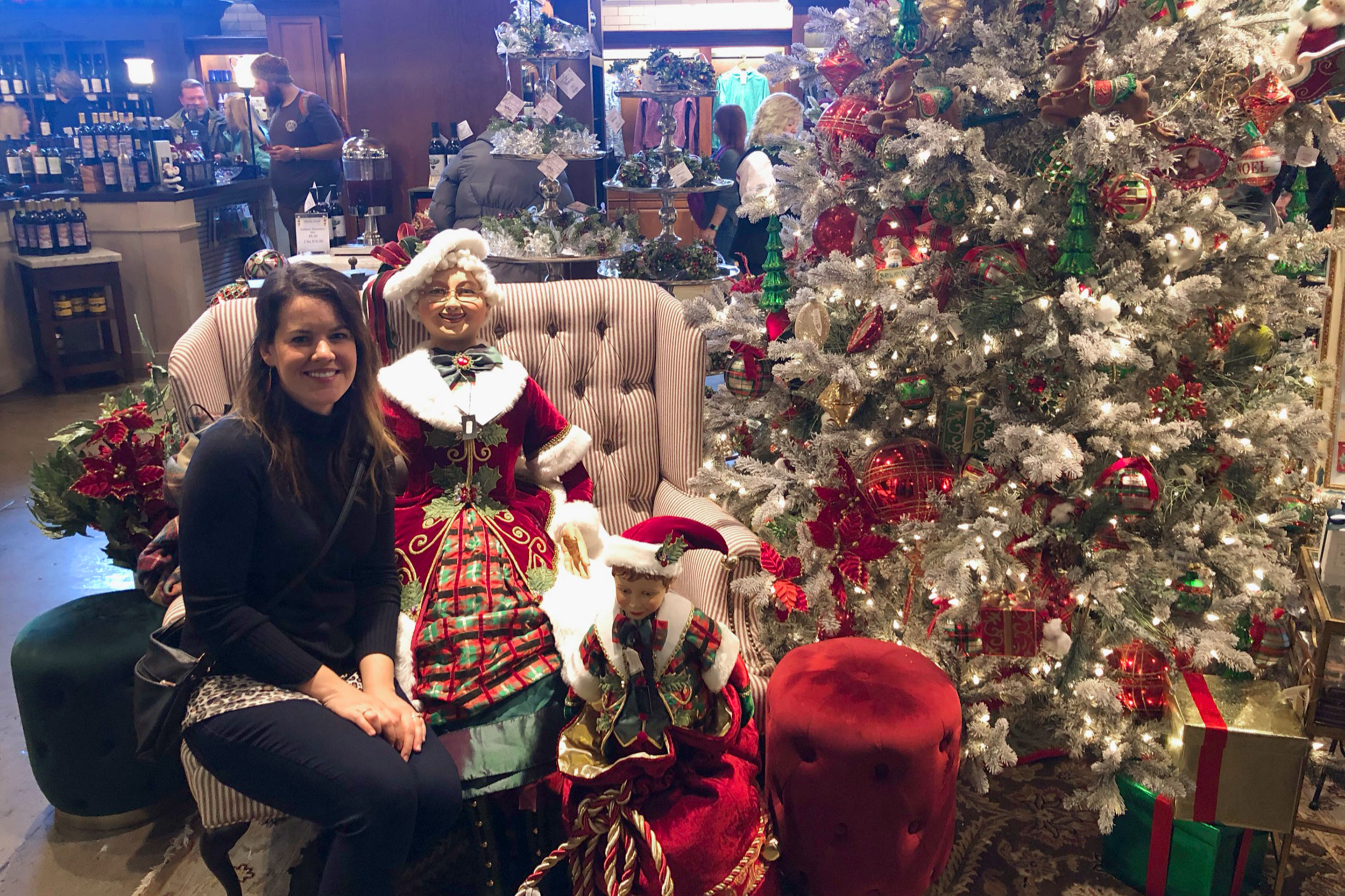 Author, Jenn C. Harmon, wears a black sweater seated near a Mrs. Claus doll near a decorated Christmas tree and other home decor items at the local Christmas Shoppe.