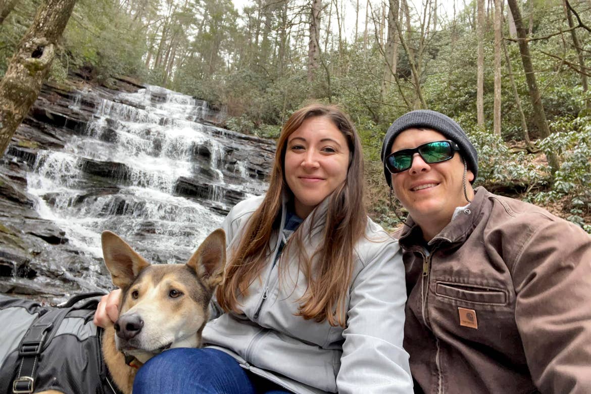 A dog, a woman in grey jacket, and a man in a brown jacket, sunglasses and a black beanie sit near the edge of a waterfall.