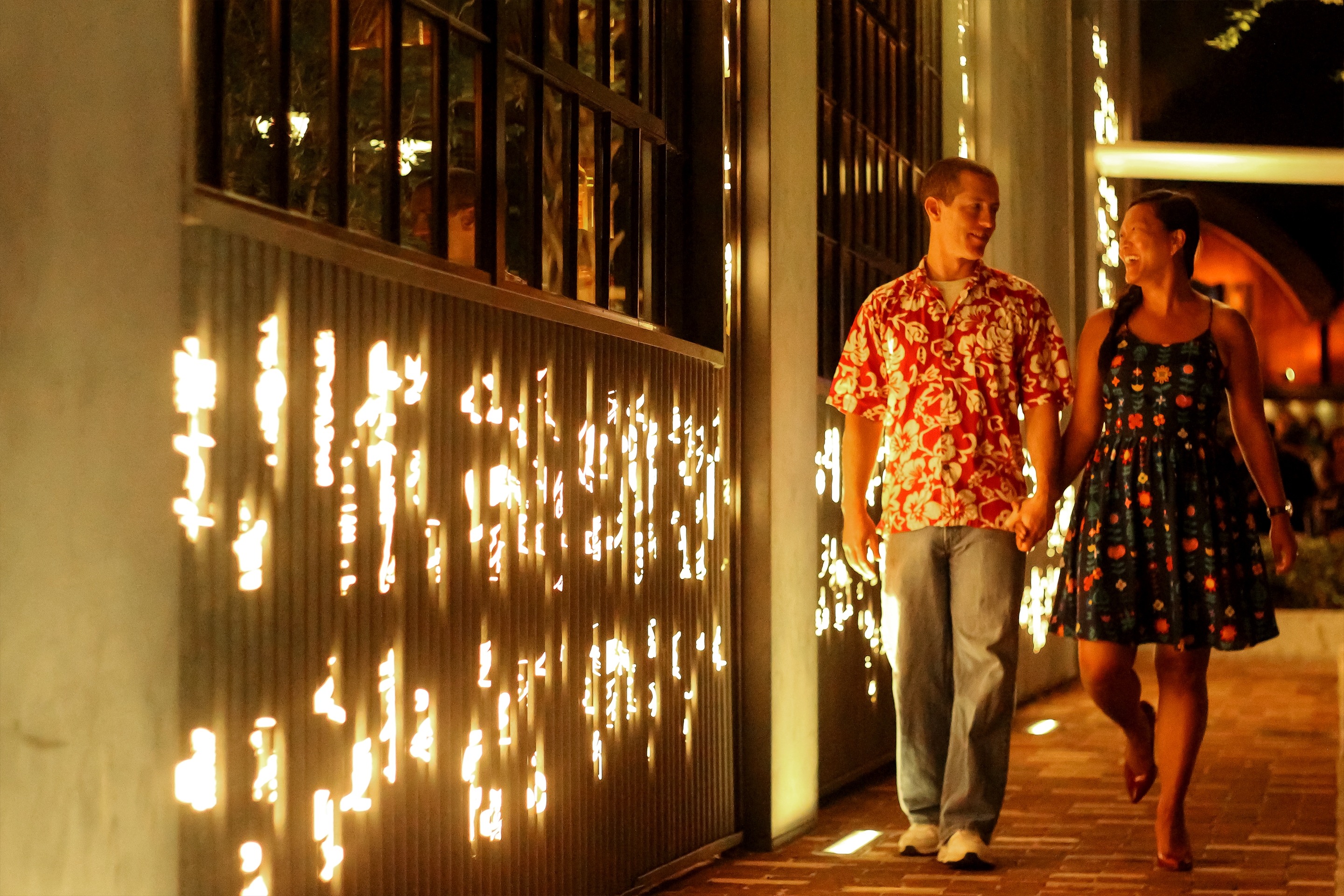 A Caucasian man wearing a red Hawaiian shirt and jeans (left) walks with an Asian woman wearing a patterned, navy dress (right) as they walk next to a building exterior with die-cut lights at night.