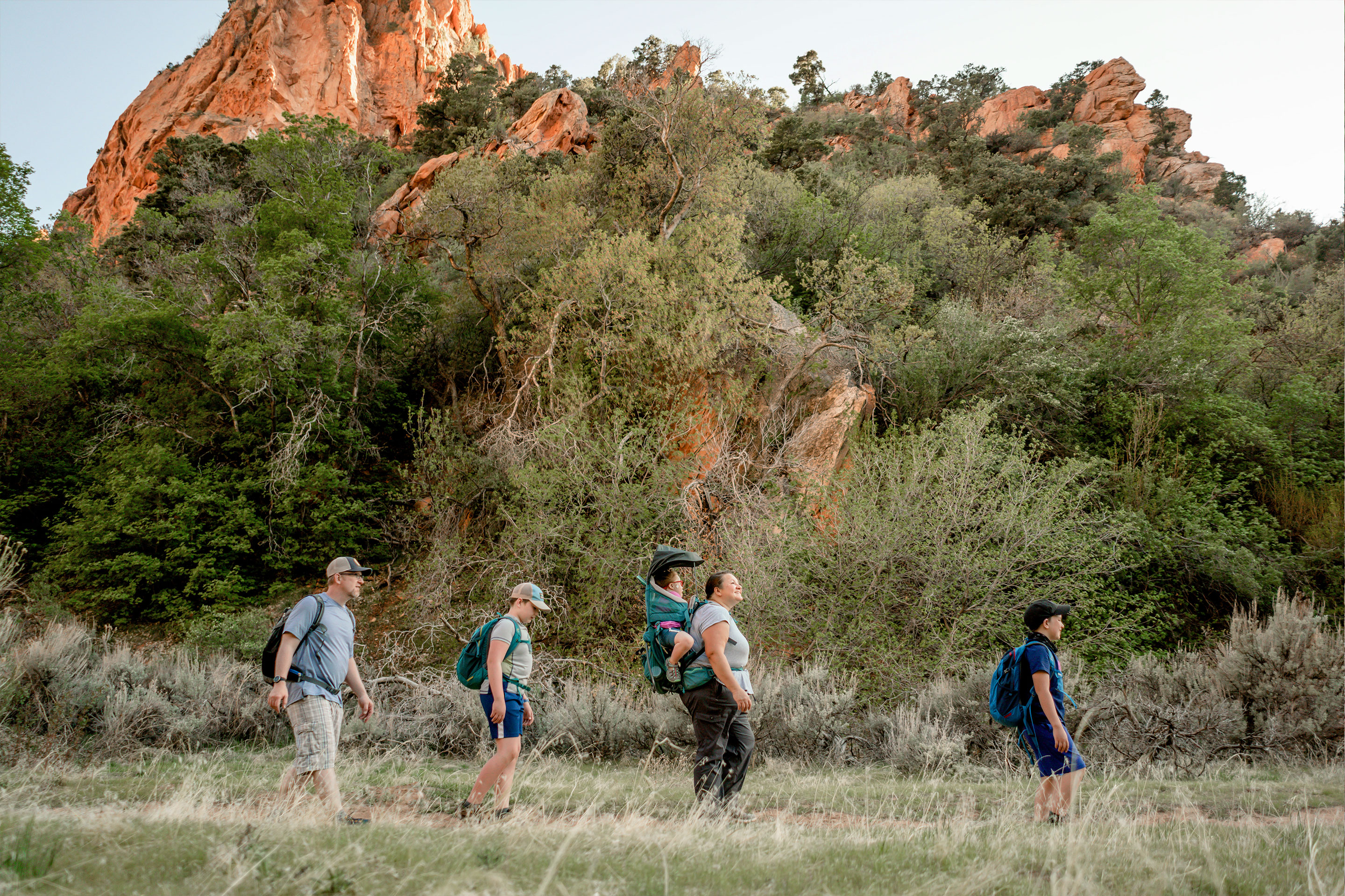 Featured Contributor, Melody Forsyth (middle-right), and her family walk single-file in front of a rock formation while wearing hiking backpacks.