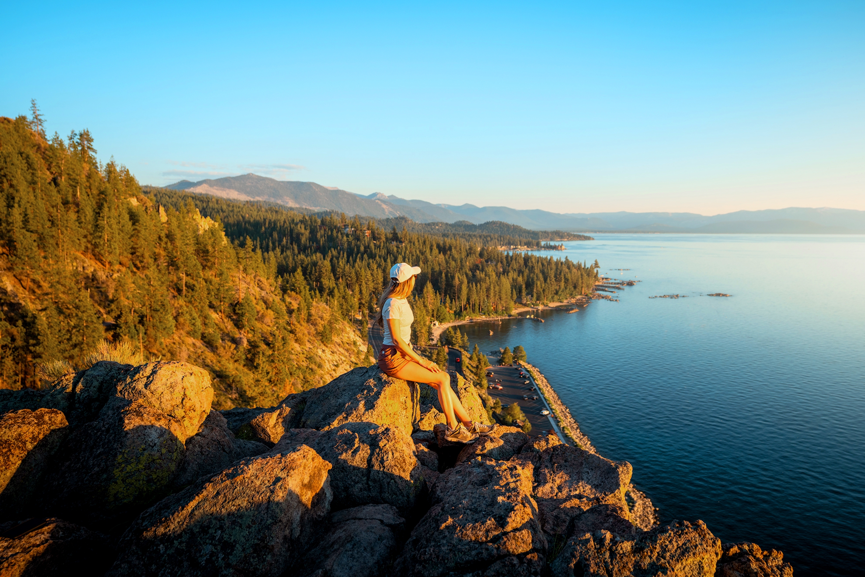 A caucasian woman wearing a white shirt, white baseball cap, shorts and boots sits on a rock overlooking a lake surrounded by pine trees on a hill.