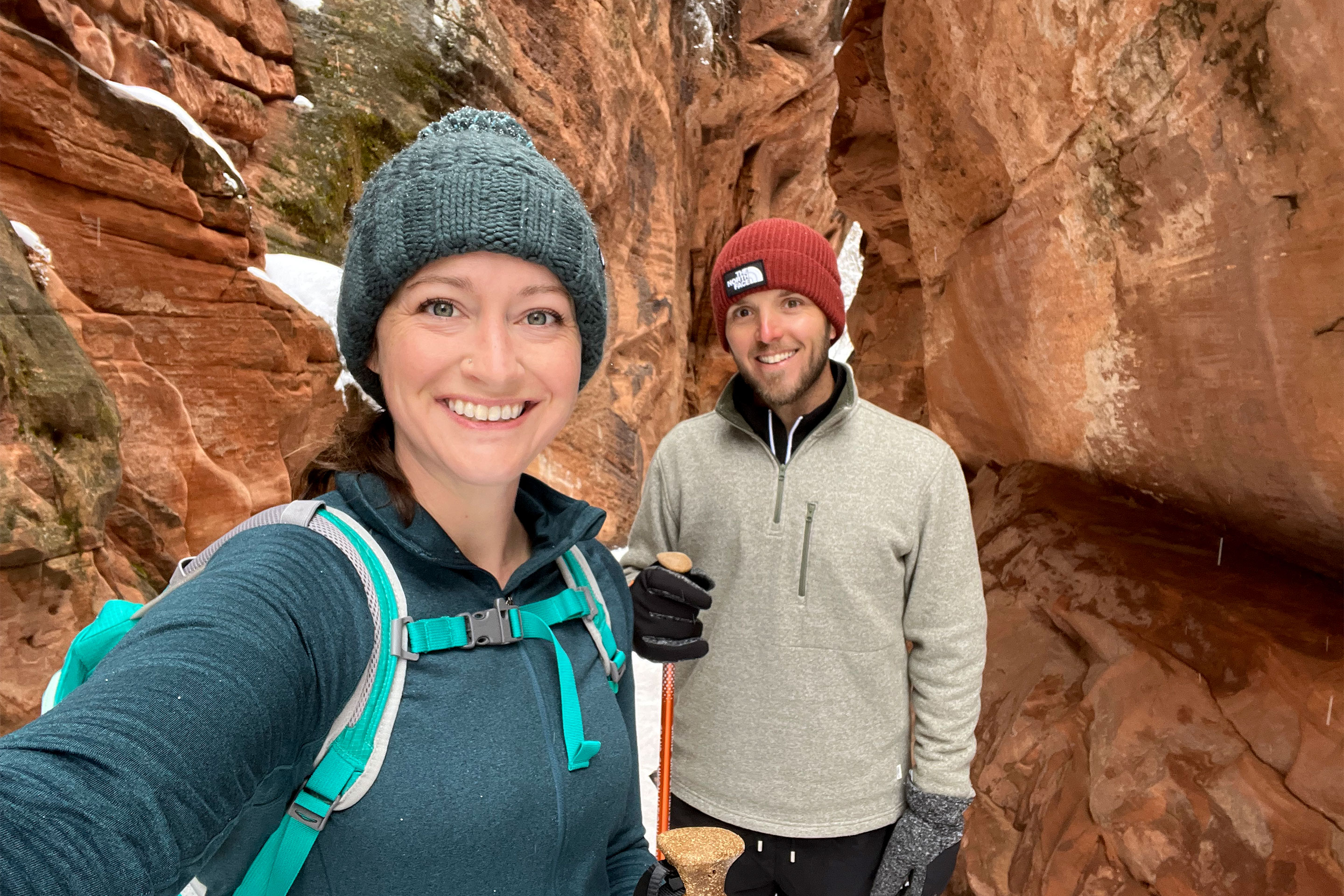 A woman in a blue light jacket, knitted hat and backpack stands next to a man in a cream pullover and red knitted hat with a walking stick in a red rock formation.