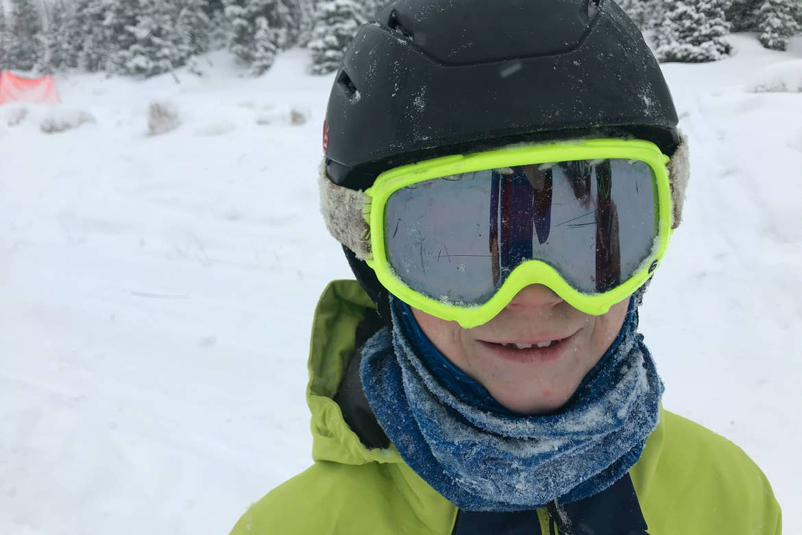 A young boy wears a black ski helmet, neon yellow goggles and winter jacket, with a blue balaclava wrapped around his neck while standing outdoors in the snow.