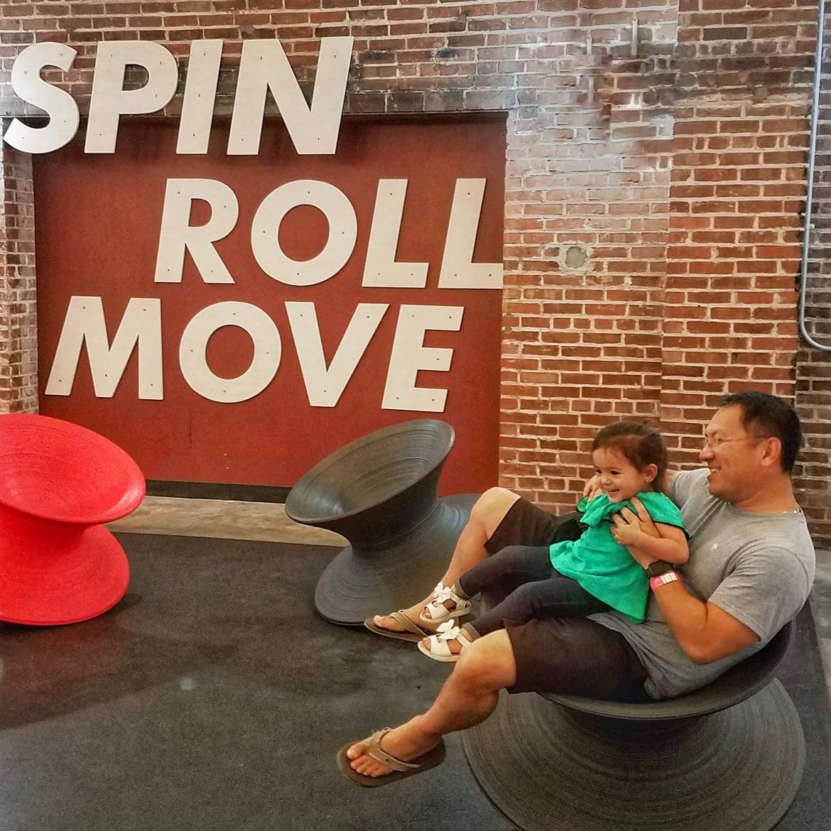A man holds a toddler on his lap while seated in a conical, black chair in a brick interior with signage that reads, 'SPIN ROLL MOVE.'