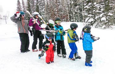 Two adults and five children wear winter apparel, ski helmets and goggle while holding a pair of ski's over their shoulders on a snowy slope.