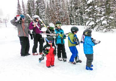 Two adults and five children wear winter apparel, ski helmets and goggle while holding a pair of ski's over their shoulders on a snowy slope.