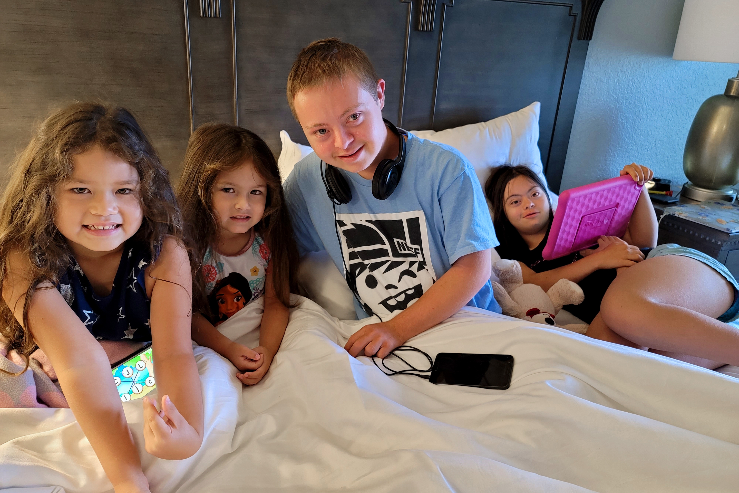 From Left to Right: Two young girls, a young boy, and another young girl holding a pink iPad sit on a large bed.