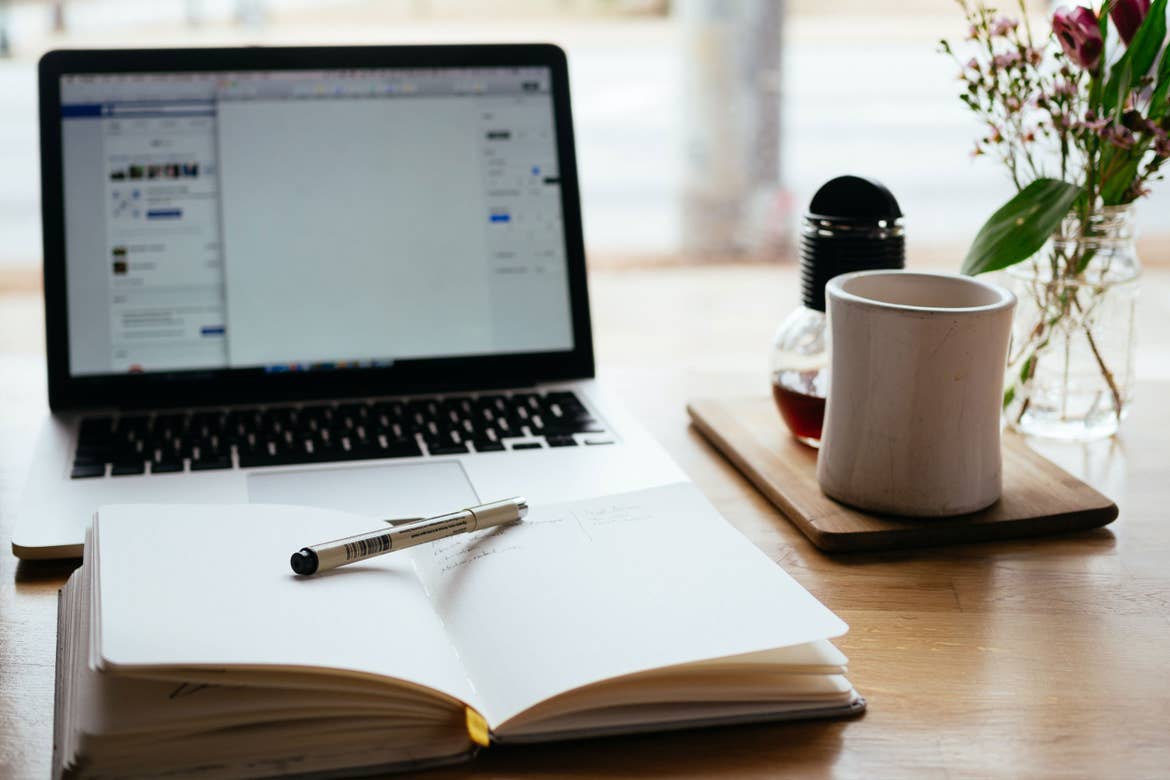 A laptop sits alongside an open notebook, coffee mug and a window.