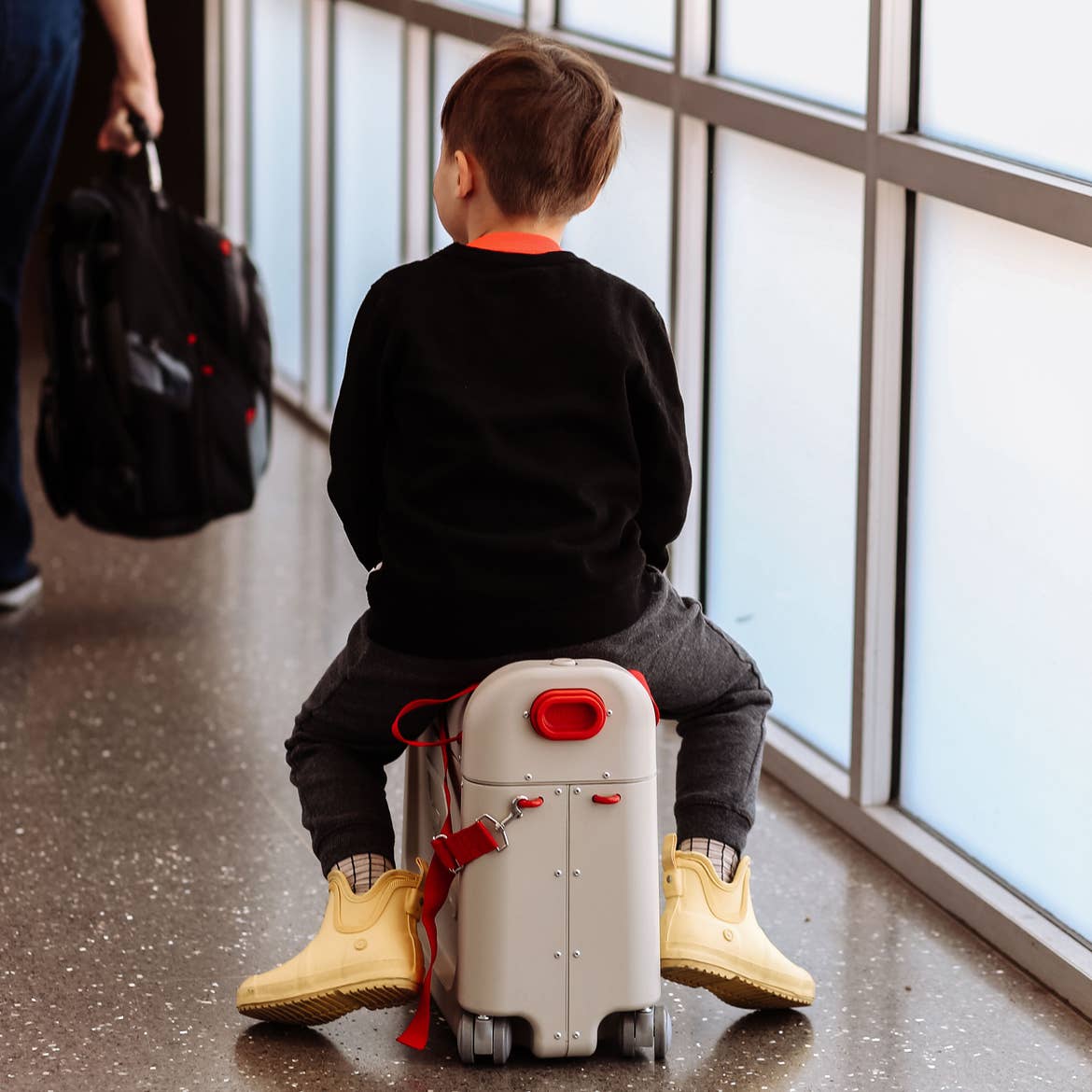 Featured contributor, Mia St. Clair's son, Grey, sits on his white wheeled suitcase in an airport terminal wearing a black shirt and yellow rain booties.